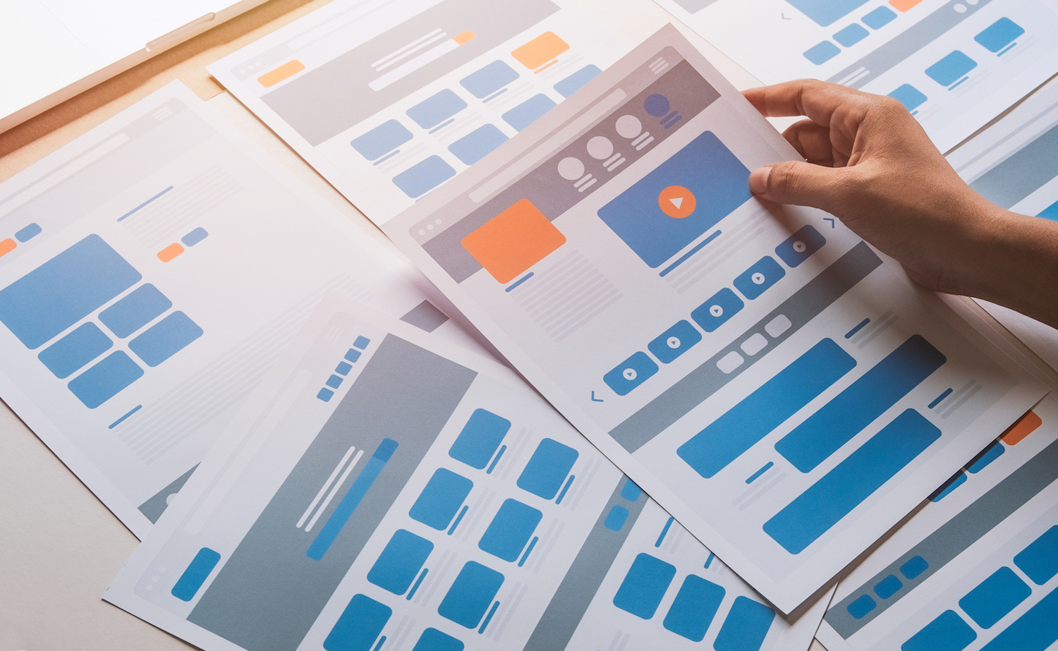 A hand holding up blue, orange, and gray wireframes of a website for usability testing.